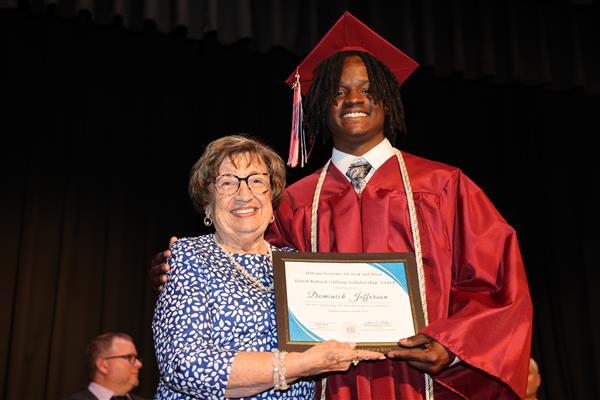 A graduate from the Alabama School for the Deaf smiles while receiving the Floyd Buford Gilliam Scholarship Award from Ms. Judith Gilliam. The graduate wears a maroon cap and gown while Ms. Gilliam wears a blue blouse with white flower pedals, a necklace, and a bracelet. 